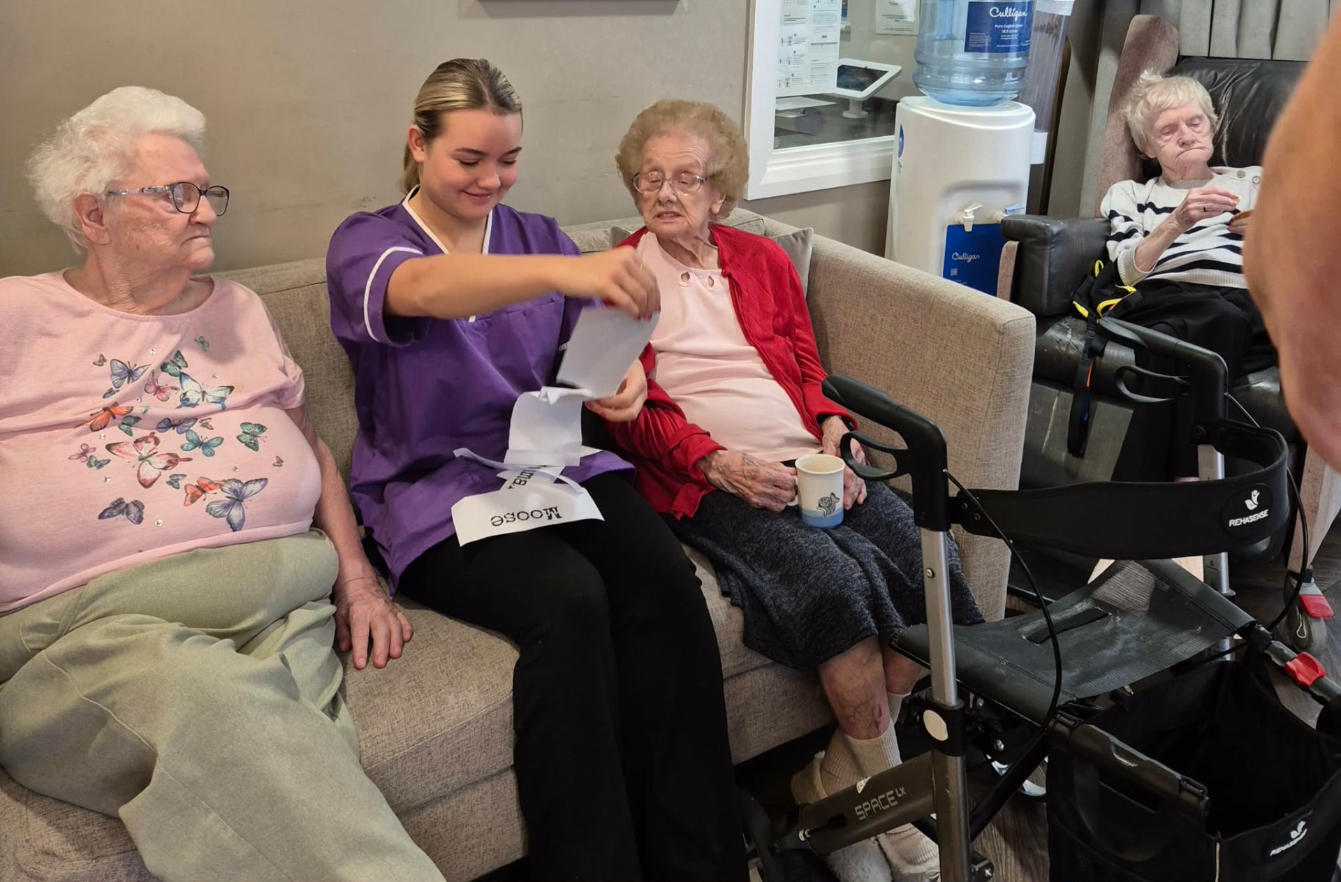 A member of care home staff running a Taskmaster task with two residents in a lounge.